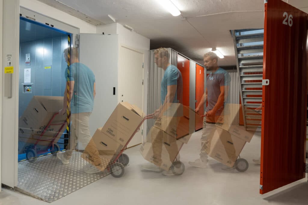 Mann som bruker en jekketralle med pappesker inn i en vareheis i et minilager, med bevegelsesslør som viser flere posisjoner.