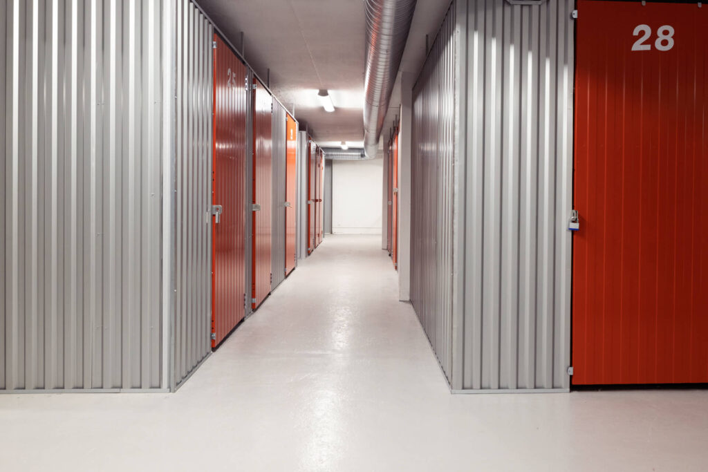 Korridor i et innendørs minilager med grå vegger og oransje dører på begge sider og et lyst gulv med jevn belysning.