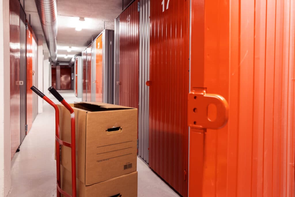 Kartongbokser på en rød tralle står i en korridor med oransje og grå lagringsboder i et innendørs minilager.