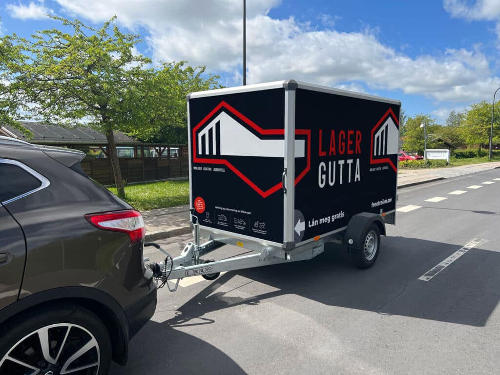 Lukket skaphenger med Lagergutta-logo koblet til mørk SUV som kjører på vei om sommeren, med norsk boligområde i bakgrunnen.